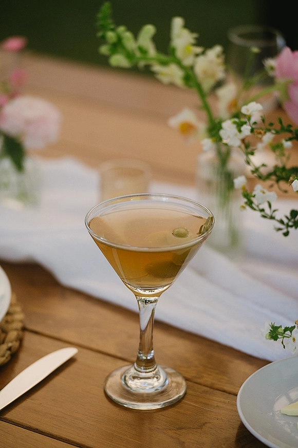 Wedding signature cocktail in a martini glass with olive garnish, set beside bud vases of white and pink flowers on a wood reception table
