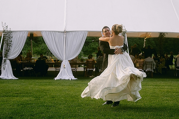 First dance as the groom holds the bride while she twirls her wedding dress under a draped tent with string lights and candles on the lawn