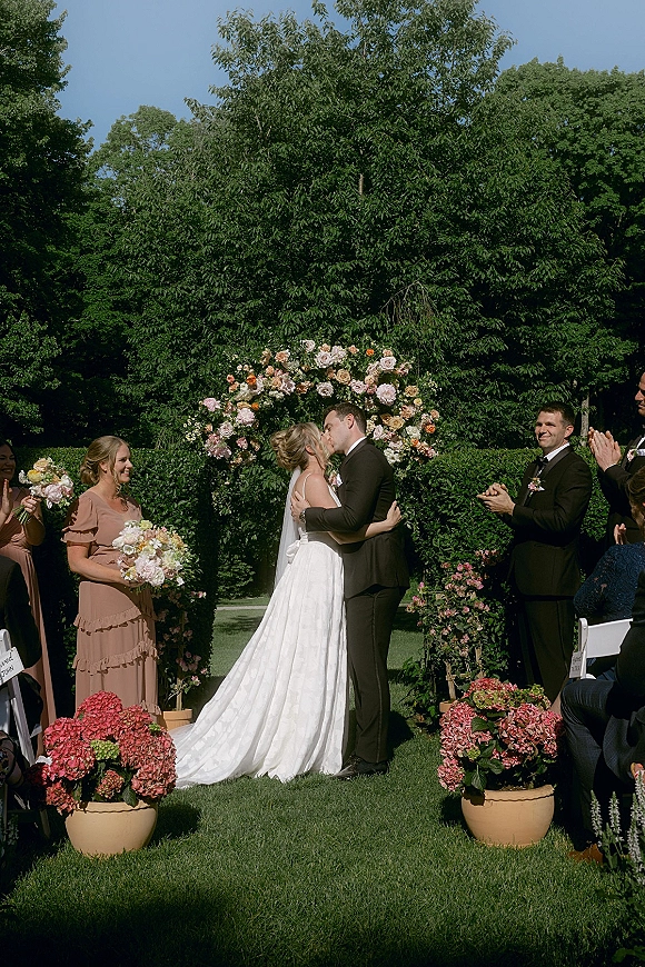 Ceremony kiss as bride and groom embrace under a round floral arch, veil flowing with bridesmaids holding bouquets on a sunny garden lawn