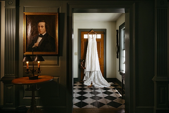 Wedding dress hanging on a wooden door with high heels below, framed portrait and wall sconces in an entryway with checkered tile floor