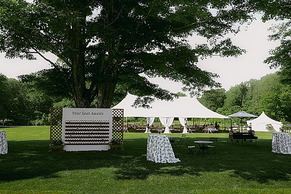 Outdoor reception setup under a sailcloth tent with cocktail tables, patterned linens, escort card display, and umbrellas on a grassy lawn