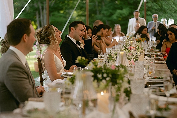 Wedding reception toast as the bride and groom laugh at a speech, seated at a long banquet table with candlelit florals under a draped tent.