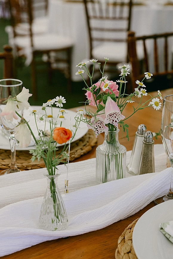 Reception tablescape with bud vase centerpiece of daisies and pink blooms beside wine glasses on a wooden table in an indoor reception space