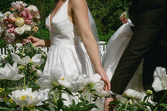 Couple portrait of bride and groom holding hands, her pink and white bouquet and veil visible, posed by garden flowers and a white fence