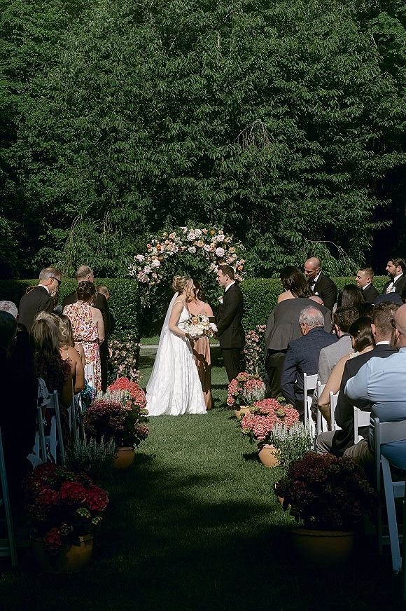 Wedding ceremony with bride and groom at altar under a floral arch, bride holding bouquet and veil flowing on a garden lawn with guests