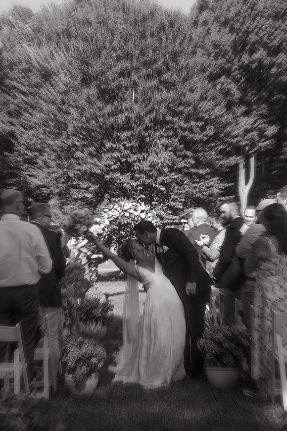 Wedding kiss moment as bride and groom share a dip kiss walking down the aisle under a floral arch, guests cheering in a garden ceremony