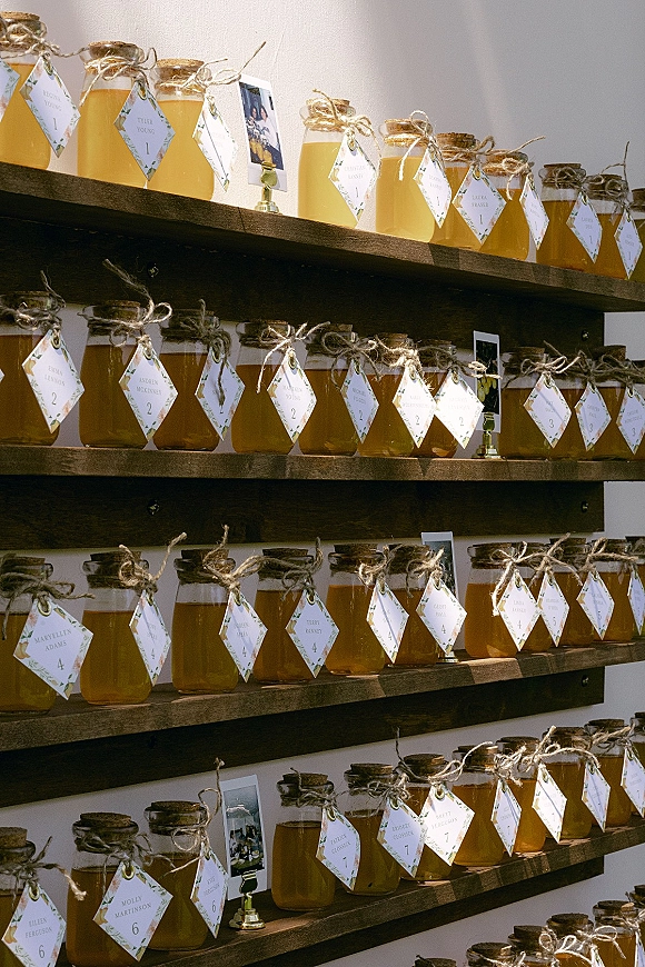 Wedding escort cards as honey jar escort cards with cork lids and twine bows, labeled tags on wooden shelves with photo clips against a white wall