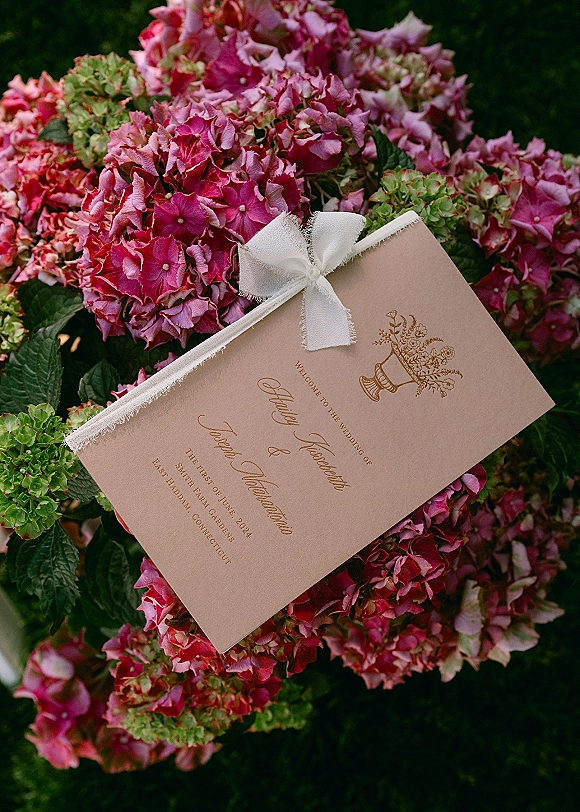 Wedding invitation with gold foil text and a deckle-edge ribbon bow, styled on hydrangea blooms amid garden greenery background