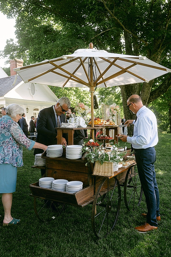 Wedding food station with tiered appetizers and fruit on a wood serving cart under a patio umbrella on an estate lawn with guests