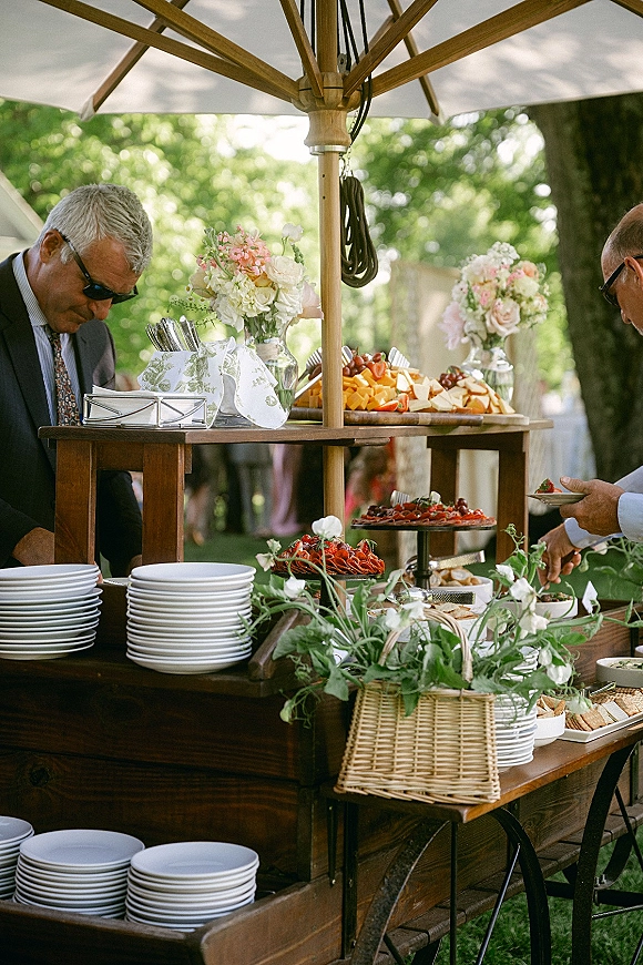 Wedding cocktail hour food display on a wood bar cart with cheese board, fruit, crackers, and florals under a patio umbrella in a garden setting