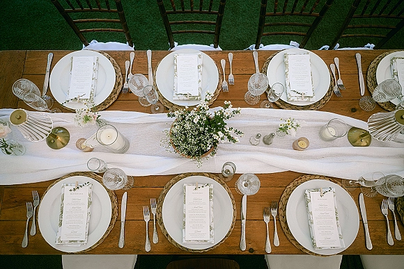 Reception tablescape with wedding place setting, woven chargers, white plates, menus, candles, and floral centerpiece on a lawn outdoors