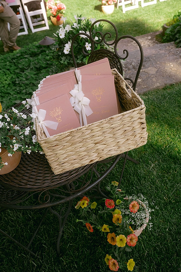 Wedding programs in blush paper with gold foil printing and ribbon bows in a wicker basket on a wrought iron chair by garden path