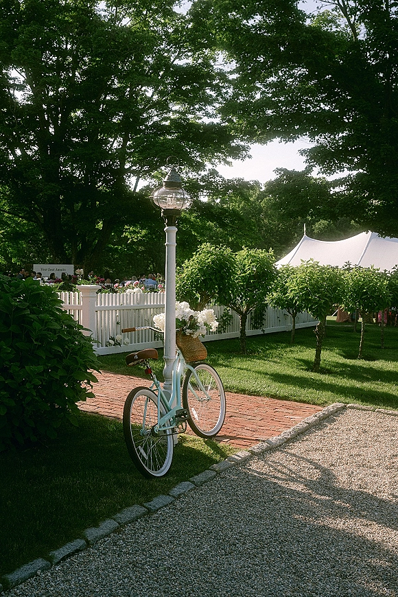 Wedding venue details with a vintage bicycle flower basket of white blooms beside a lamppost and picket fence along a brick path