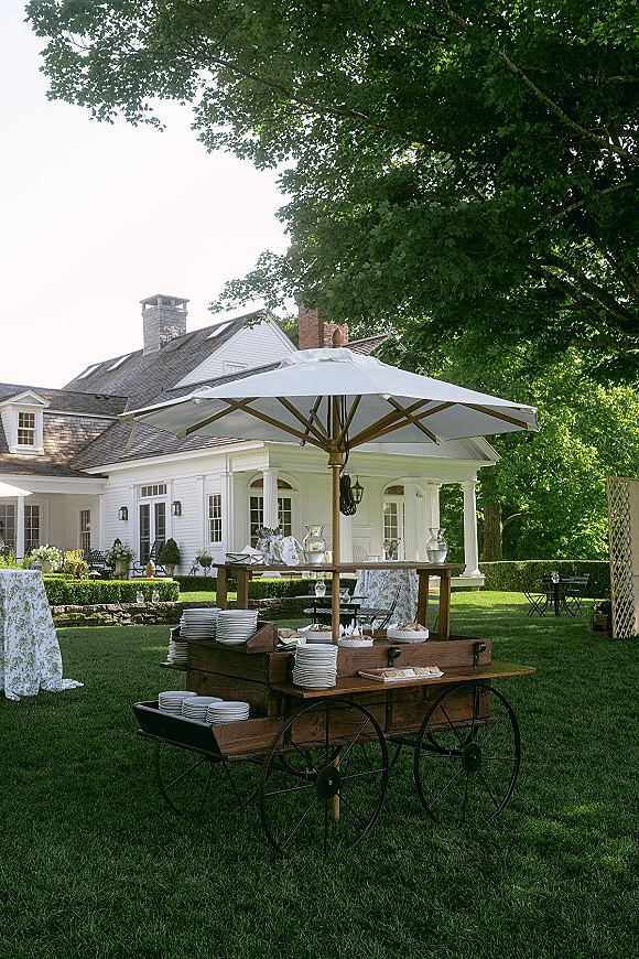 Outdoor drink station with glass beverage dispensers on a wood bar cart, under a patio umbrella on a green lawn by a white house