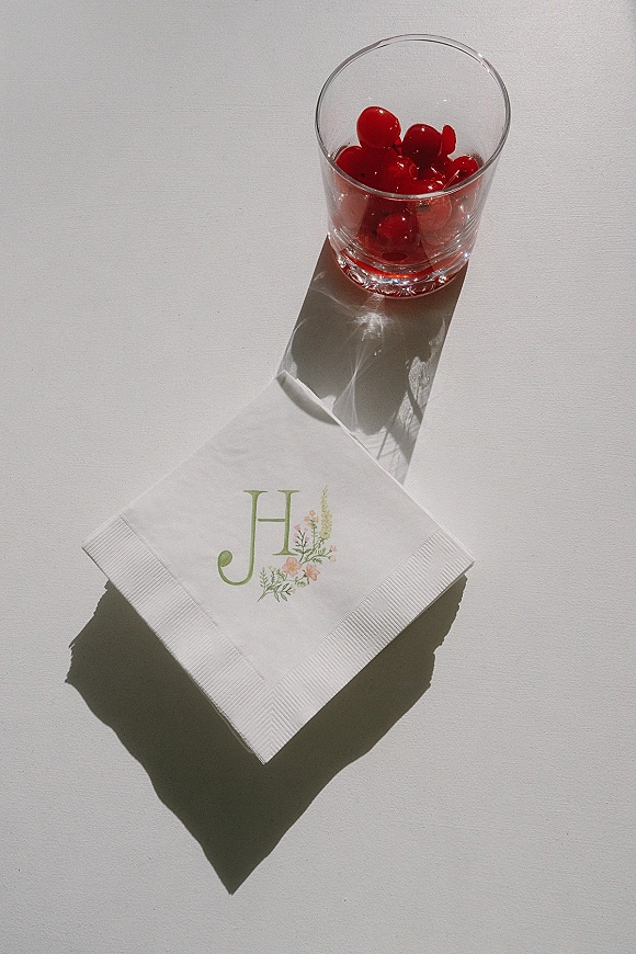 Wedding cocktail napkin with personalized cocktail napkins monogram, floral illustration, and glass tumbler with maraschino cherries on white tabletop