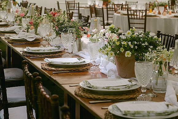 Reception tablescape on a wood banquet table setting with rattan chargers, garden florals, taper candles, and soft table lamps in a dining room