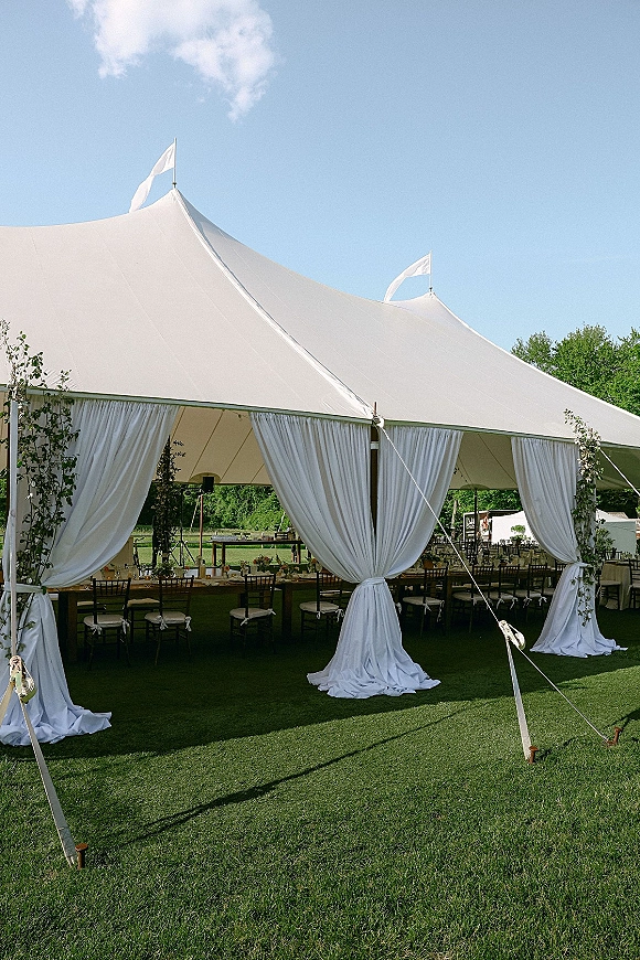 Reception tent decor in a sailcloth wedding tent with white draped curtains, greenery garlands, long tables and chiavari chairs on a sunny lawn