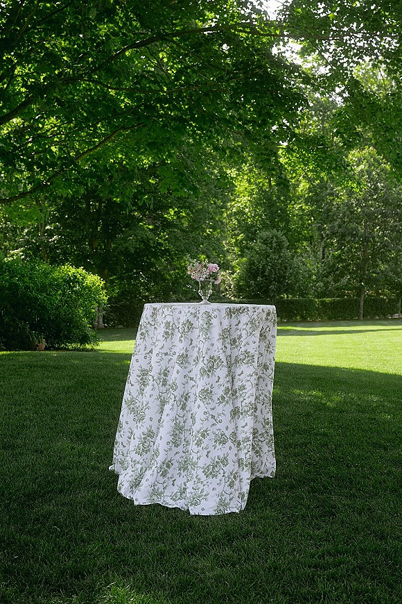 Cocktail table decor on a wedding cocktail table with floral linen cloth and silver pedestal vase of pastel blooms on a sunny lawn