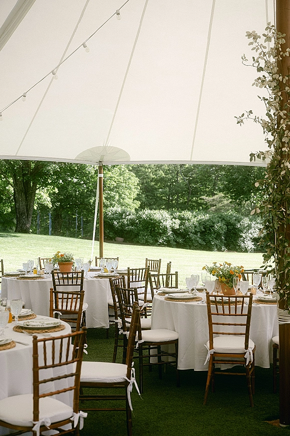 Reception tablescape under an outdoor tented reception with round tables, white linens, wicker chargers, terracotta flowers, and string lights on a garden lawn