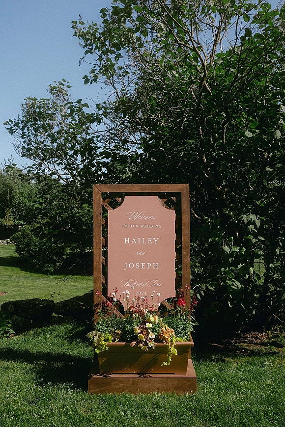 Wedding welcome sign in a wood frame with blush panel and floral planter box, set on a green lawn with trees and blue sky
