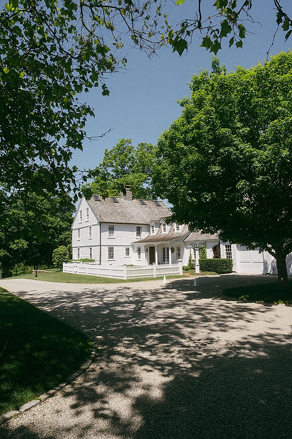Wedding venue exterior with a white clapboard farmhouse, picket fence and gravel driveway, shaded by trees under a bright blue sky