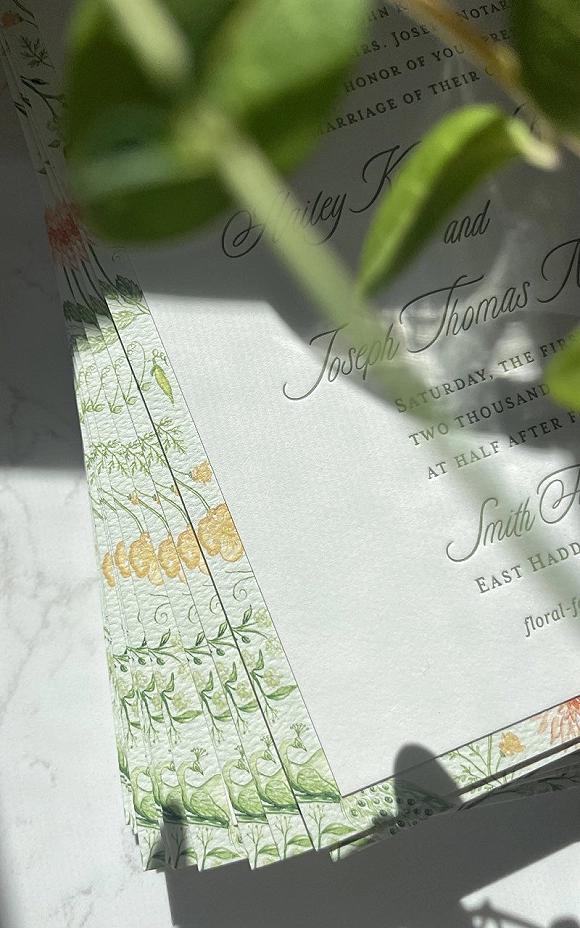 Wedding invitation suite with botanical border illustration and calligraphy, stacked on white marble with soft shadows and green leaves