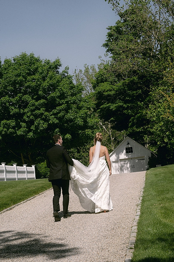 Couple portrait of newlyweds walking away, bride lifting her wedding dress train and veil beside a barn on a gravel driveway
