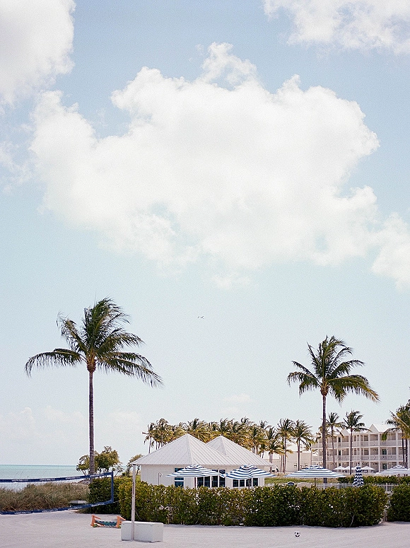 Beach wedding venue with destination wedding venue vibes, cabana tent and striped beach umbrellas on sandy shore by ocean and palms