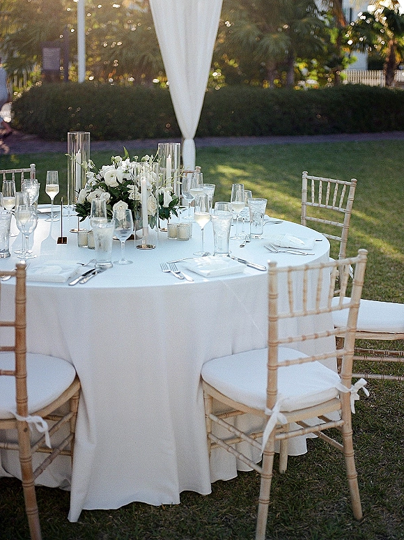 Reception tablescape with an outdoor reception table draped in white linen, white florals and greenery, cylinder vases with floating candles, and Chiavari chairs on a lawn backdrop