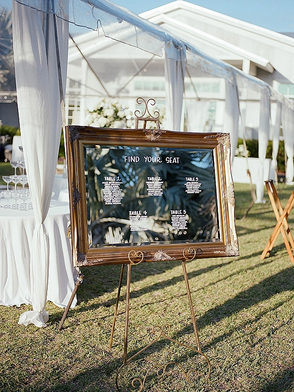 Wedding seating chart on a gold ornate framed mirror sign on an easel, beside a cocktail table with champagne coupes under a clear-top tent on lawn