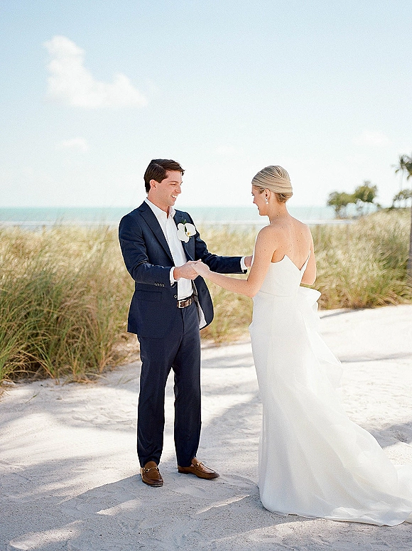 First look moment at a beach wedding first look as bride and groom hold hands on sandy dunes with ocean and blue sky behind