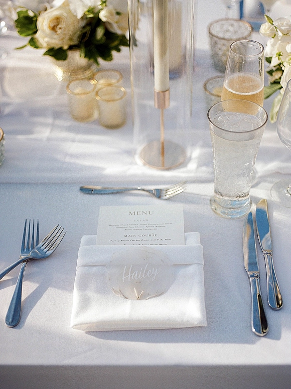 Reception place setting with white linen napkin, printed menu and place card, silver flatware, glassware, and candles amid white roses greenery