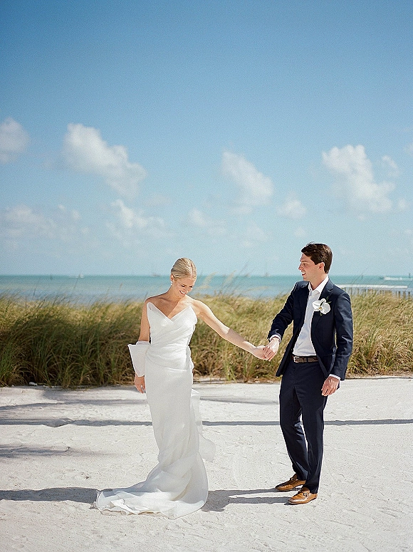 Couple portrait of bride and groom holding hands, her strapless dress train flowing as they walk a sandy beach with dunes and ocean horizon