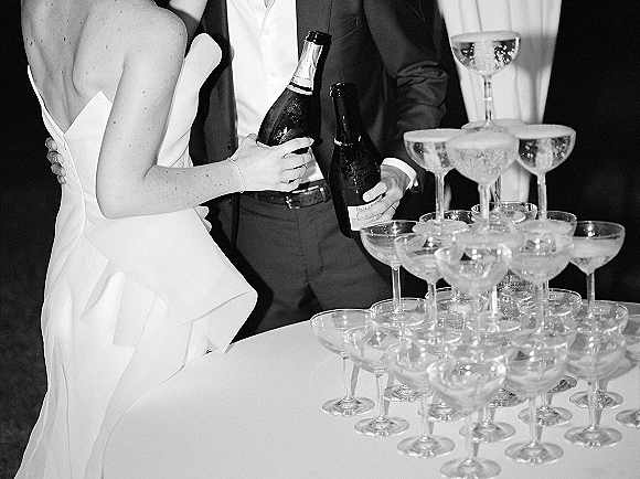 Champagne tower setup with champagne coupe tower of stacked glasses as bride and groom in dress and suit pour beside draped reception table
