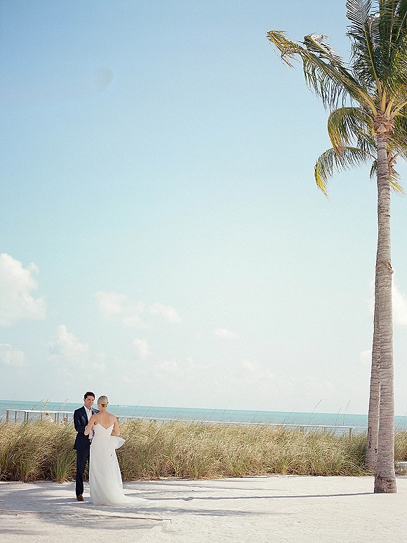 Couple portrait on a beach wedding portrait, bride in a strapless gown and groom in a suit holding hands by the ocean and palm trees