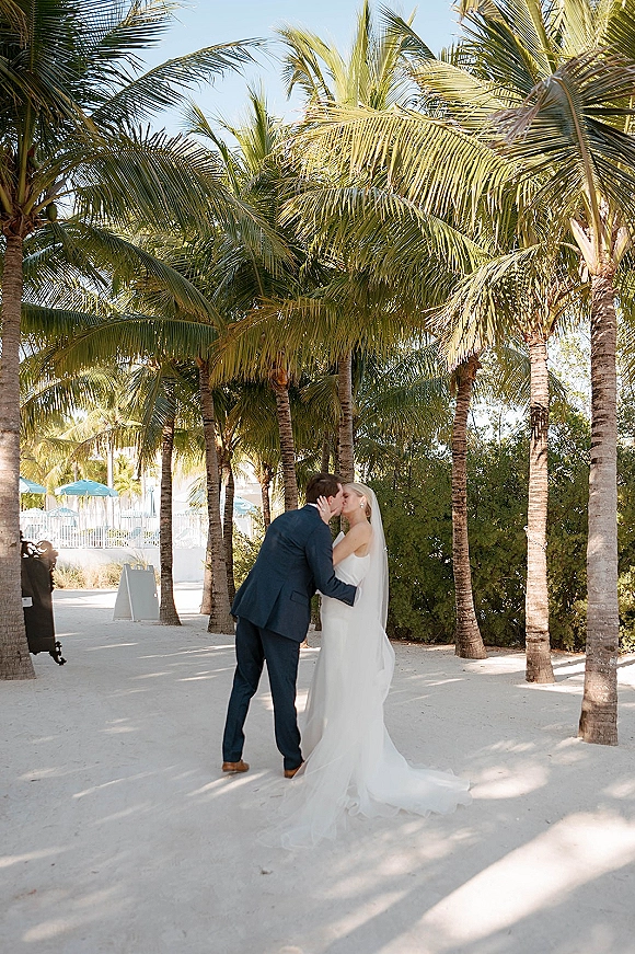 Wedding kiss portrait of bride and groom kissing, her veil flowing beside his navy suit on a sandy palm-lined path under blue sky
