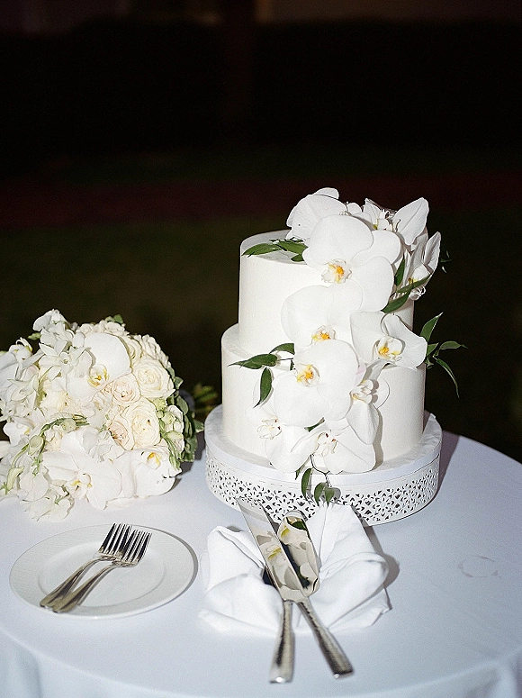 Wedding cake with buttercream frosting, three tiers topped with cascading white orchids, on a stand beside a white bouquet against dark greenery