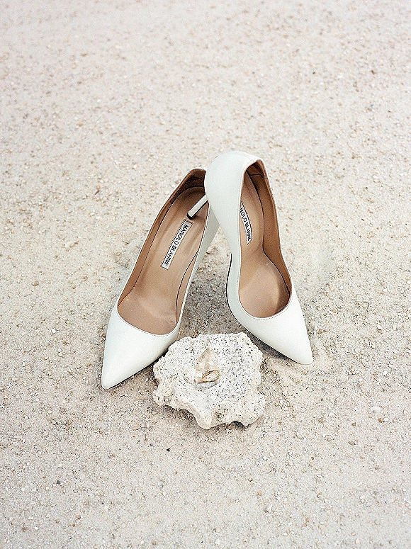 Bridal shoes in a wedding shoe flatlay with white pointed toe heels and wedding rings on a stone against soft sand background