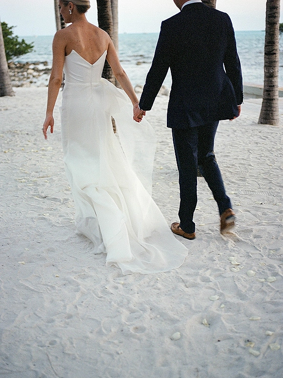 Couple walking away, bride and groom holding hands on a sandy beach with ocean water and palm trees, her strapless dress train trailing