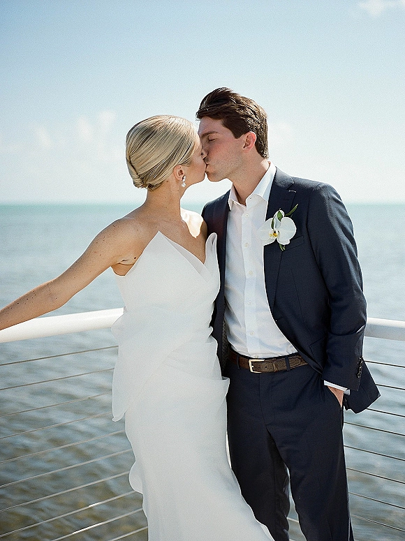 Wedding kiss portrait of bride and groom kissing, strapless gown and navy suit with orchid boutonniere on a balcony above the ocean