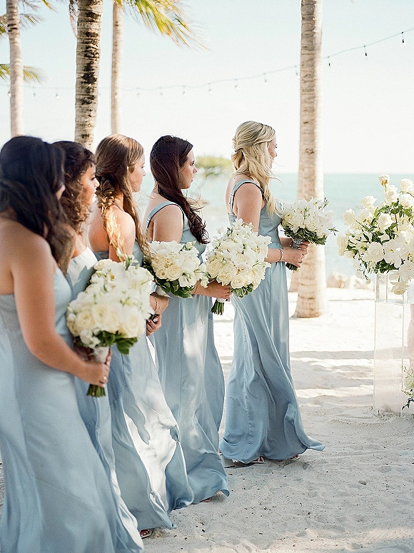 Bridesmaids ceremony lineup in dusty blue dresses holding white rose bouquets on a sandy beach with ocean, palm trees, and string lights