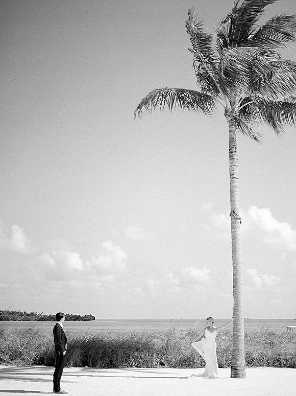 Couple portrait on a beach wedding portrait with bride holding her dress and veil beside groom in suit, palm tree and ocean behind