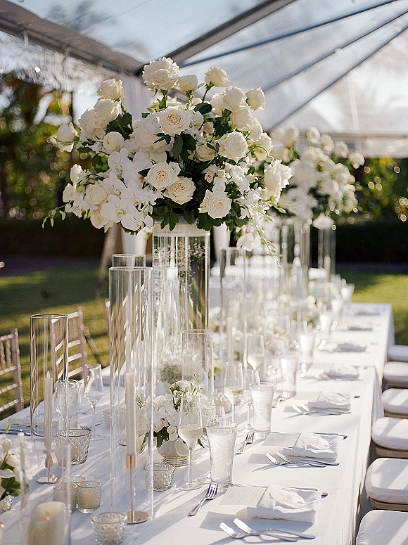 Reception tablescape with long head table decor, white rose centerpieces in tall glass vases and candlelight under a clear tent canopy outdoors