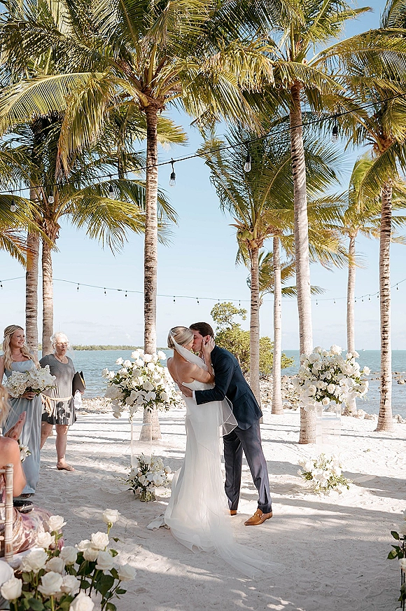 Wedding kiss at a beach wedding ceremony, bride’s veil blowing as palm trees and ocean frame the sandy aisle under string lights