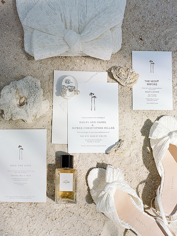 Invitation suite wedding invitation flat lay on sand with envelopes, save the date and rehearsal dinner cards, ring, earrings, perfume, seashells