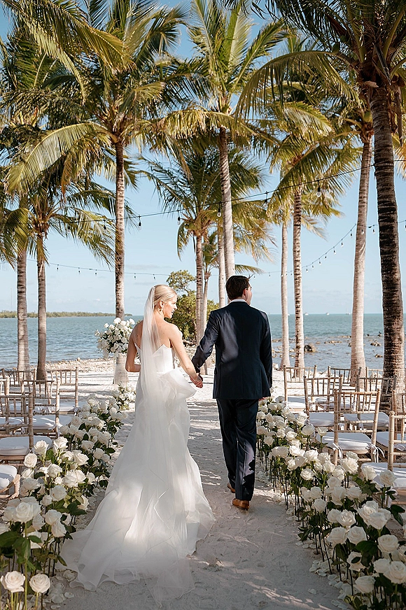 Wedding recessional as bride and groom walk away holding hands down a rose-lined aisle on a sandy beach with palm trees and ocean backdrop