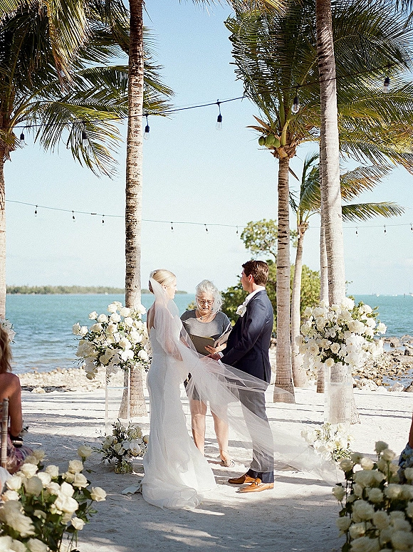 Wedding ceremony moment on the beach as bride and groom face the officiant, long veil in the breeze under string lights by the ocean
