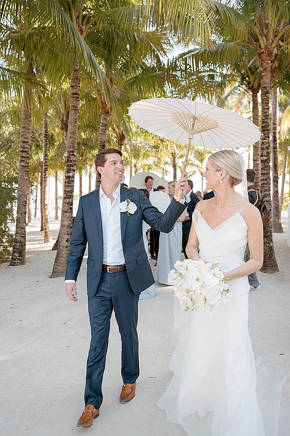 Couple portrait of bride and groom walking under a white parasol, bouquet in hand, along a sandy beach path with palm trees and ocean behind