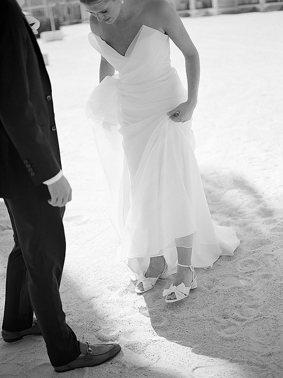 Bride portrait in a black and white wedding photo, looking down while adjusting her strapless dress with a tulle overskirt on a bright sand beach