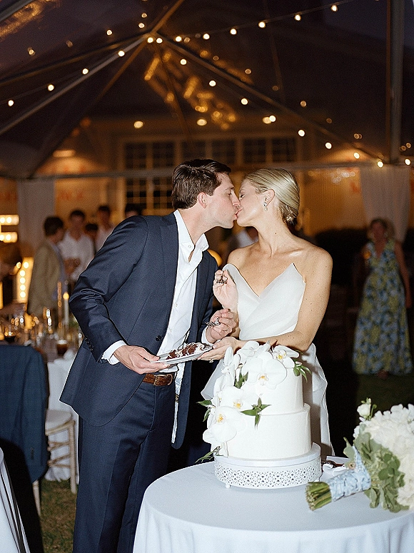 Wedding cake cutting as bride in strapless dress and groom in navy suit slice an orchid-topped cake under string lights in a tent reception